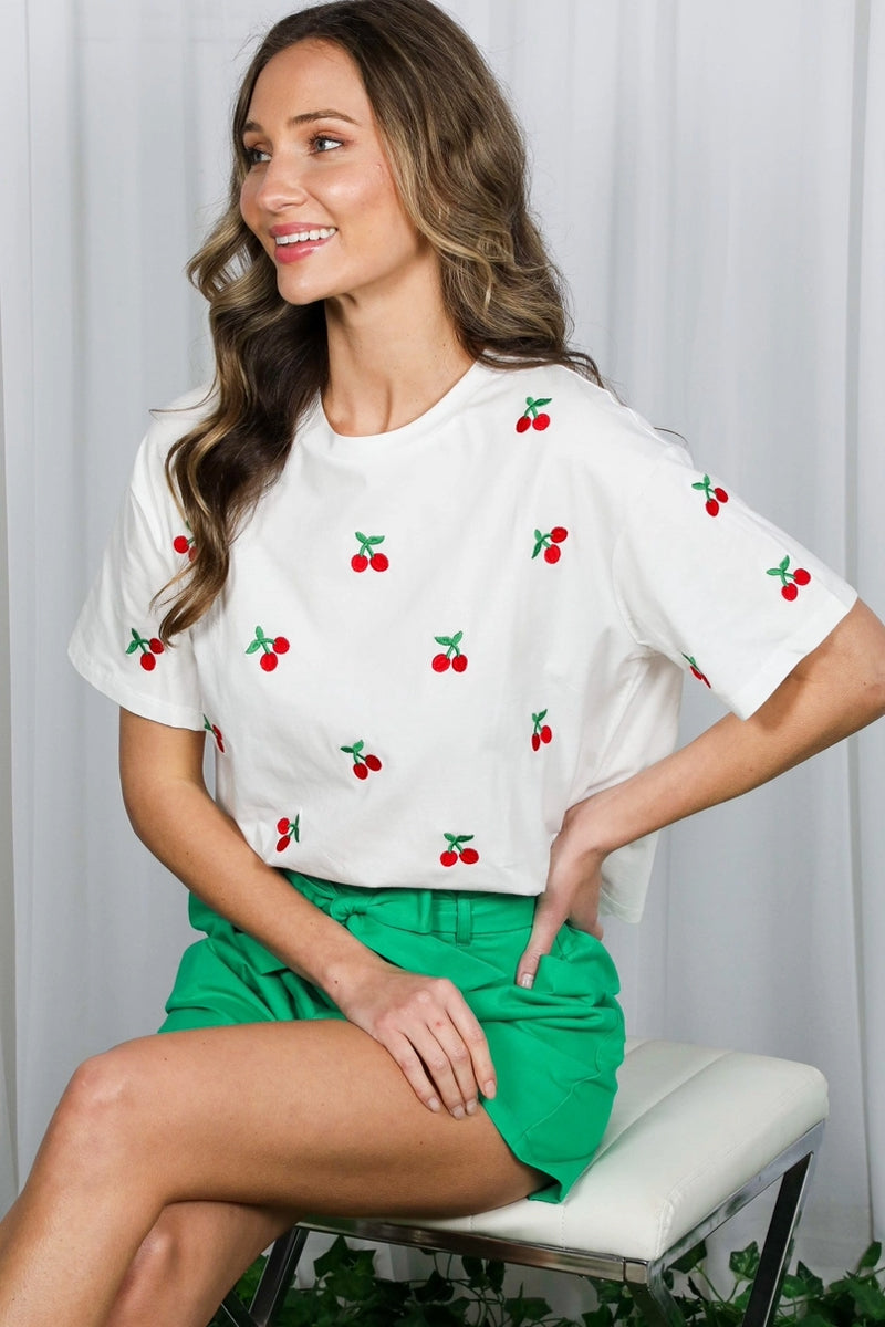 Woman wearing a white t-shirt with cherry embroidery and green shorts, sitting on a white stool.