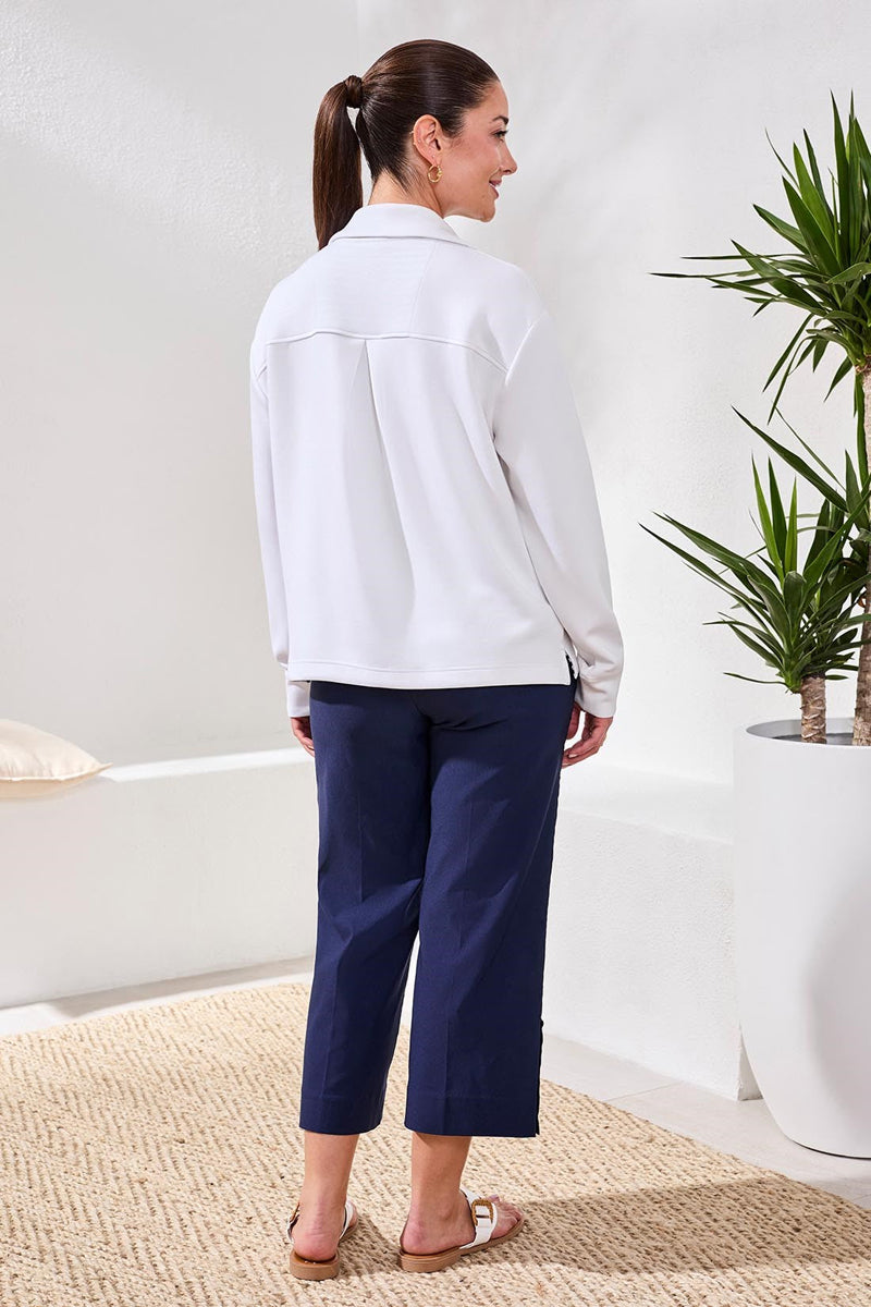 Woman wearing a white top and navy pants standing in a room with a plant.