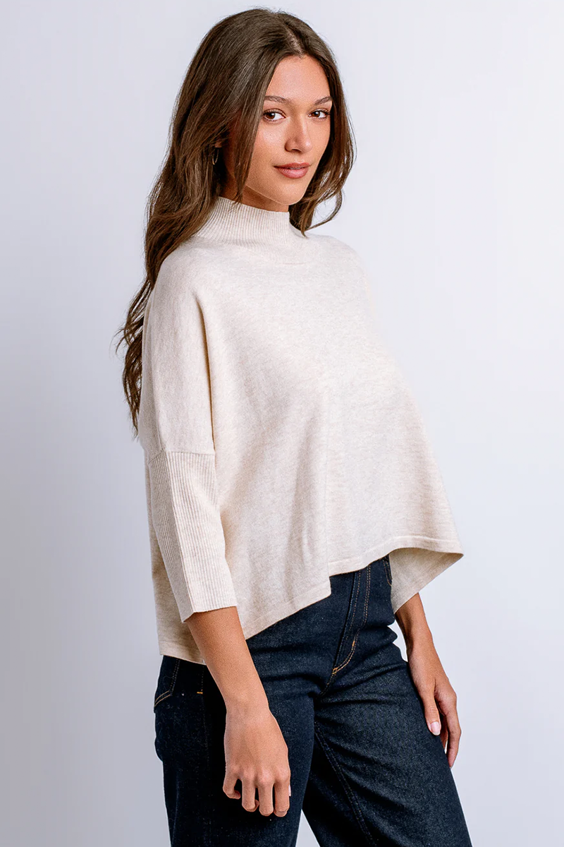 Woman wearing a beige mock neck sweater over dark jeans on a white background
