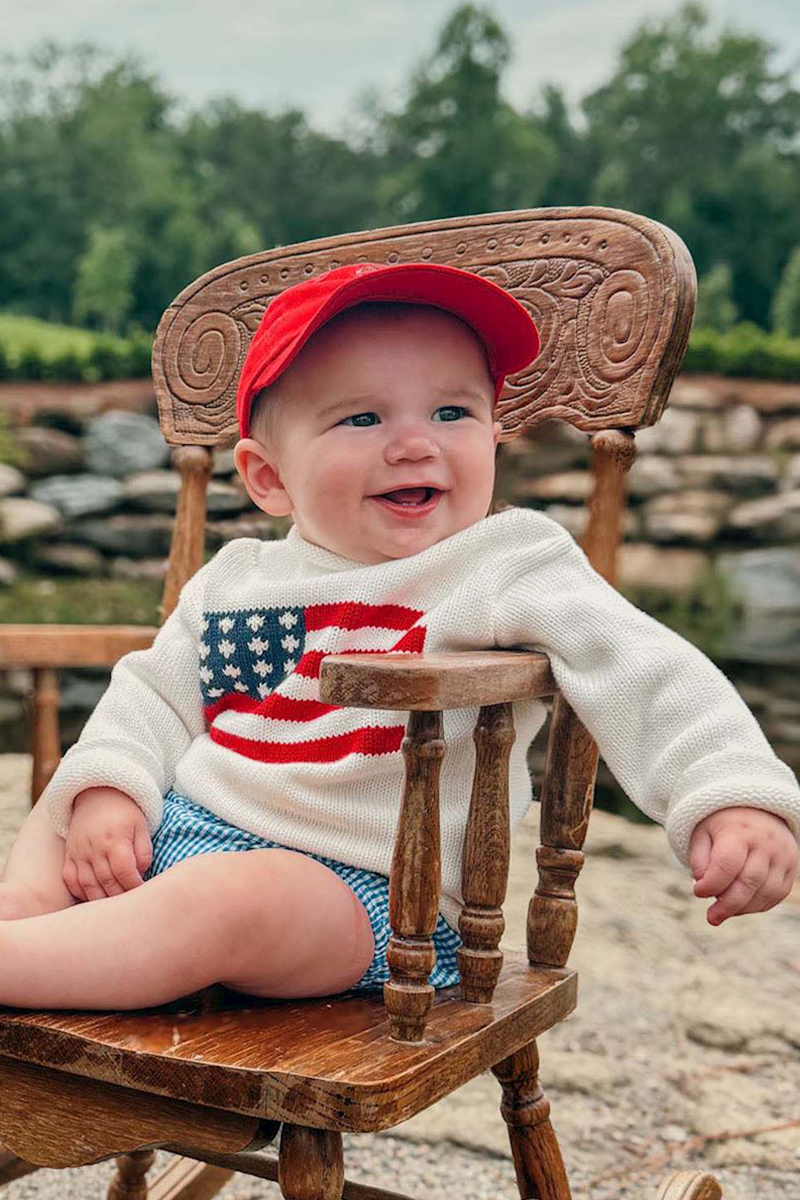 Baby sitting on a wooden chair wearing a red cap and a sweater with an American flag design.