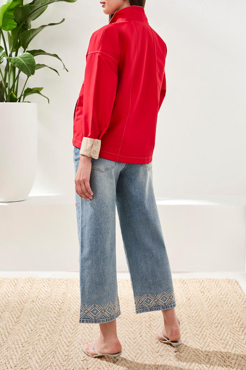 Person wearing a red jacket and blue jeans standing indoors with a plant in the background.