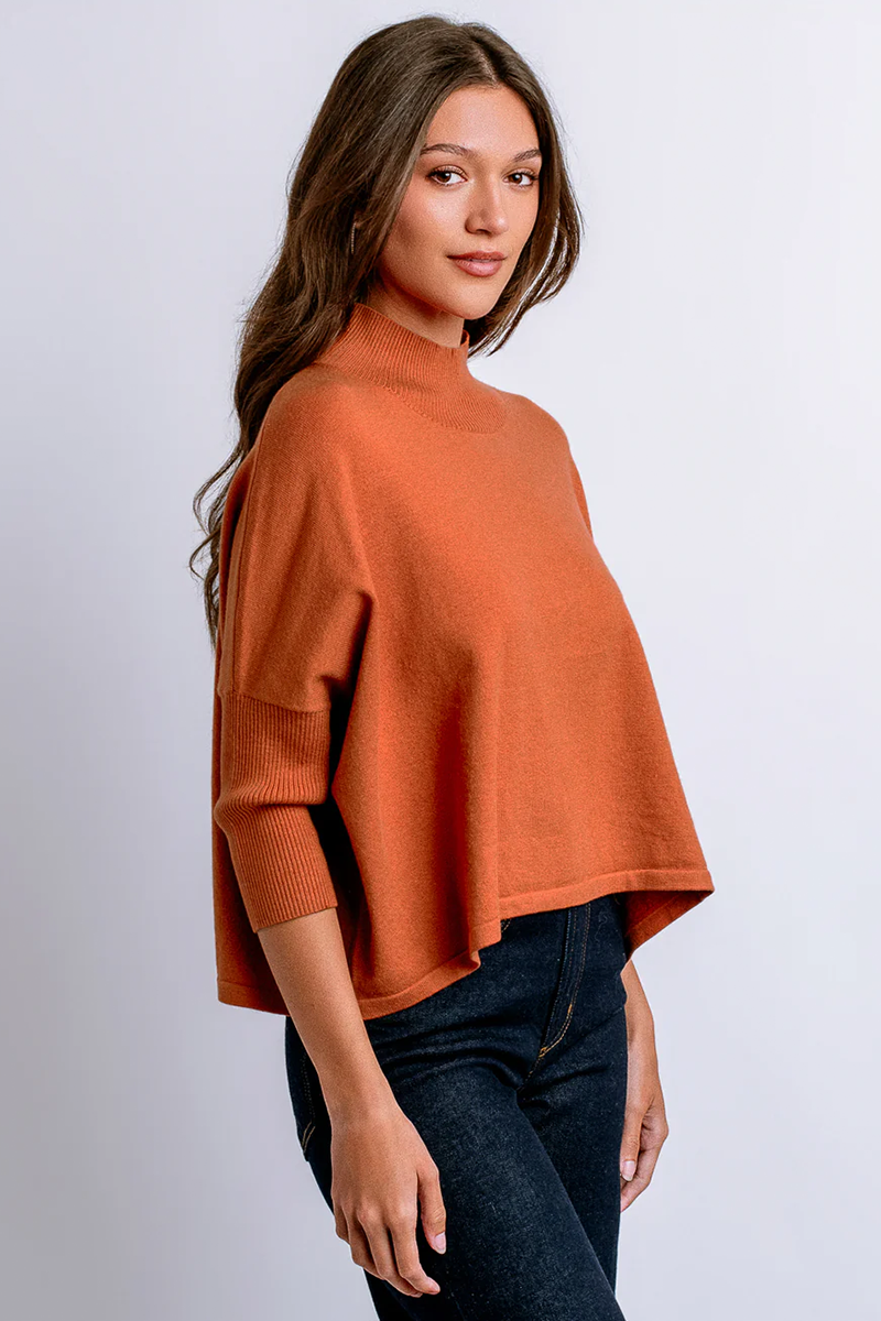 Woman wearing an orange mock neck sweater and dark jeans against a white background