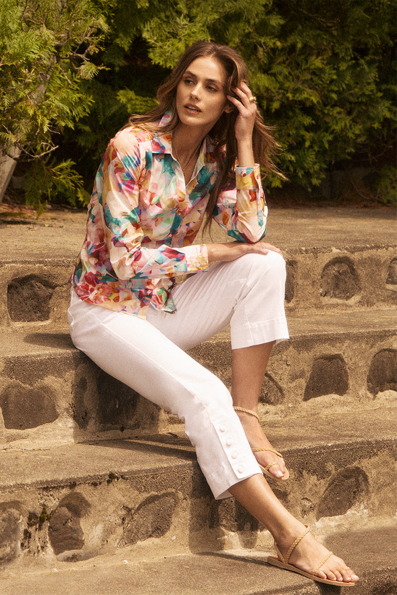 Woman sitting on stone steps wearing a colorful floral blouse and white pants.