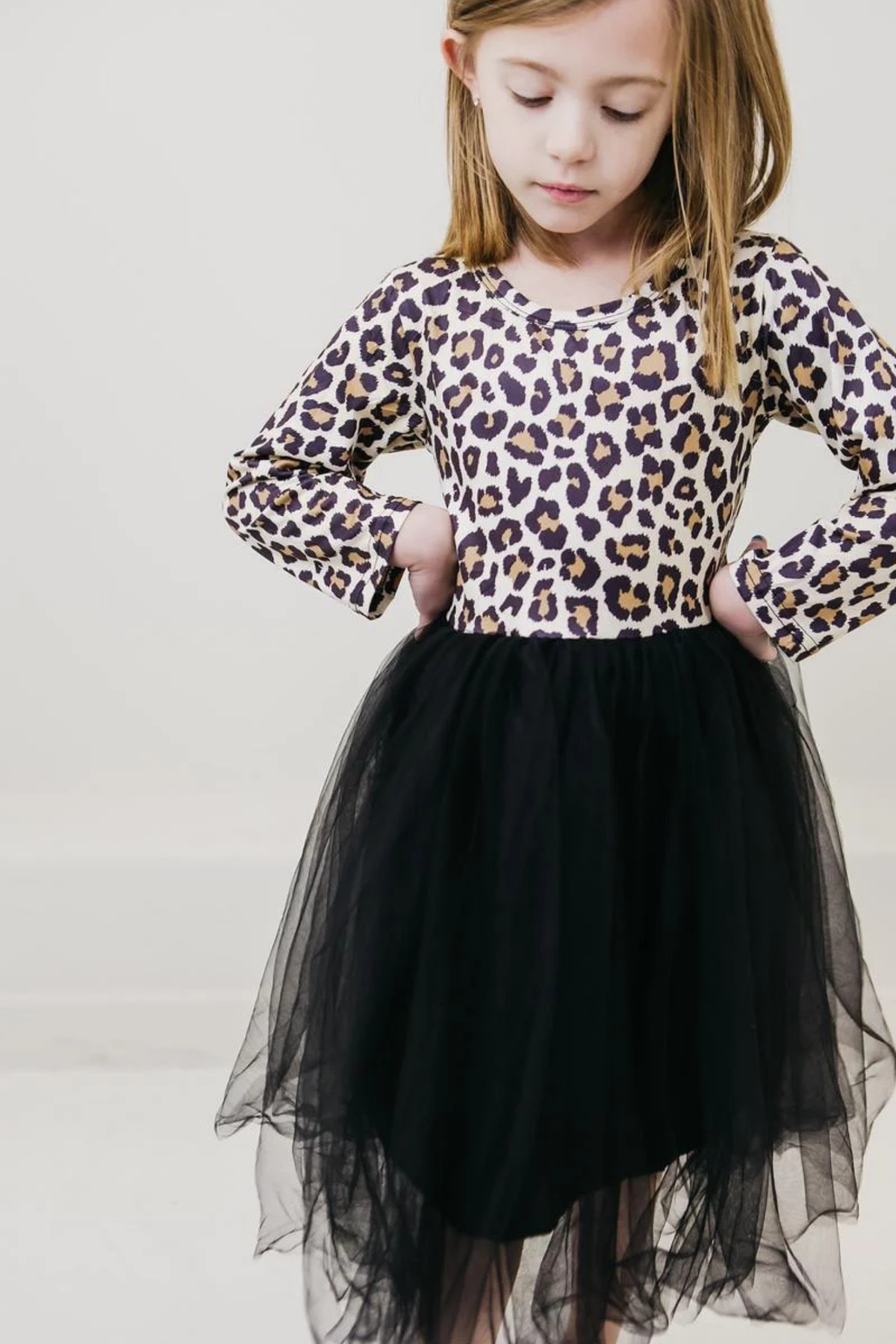 A young girl wearing a long-sleeve leopard print top and a black tulle tutu skirt, standing against a plain light background.