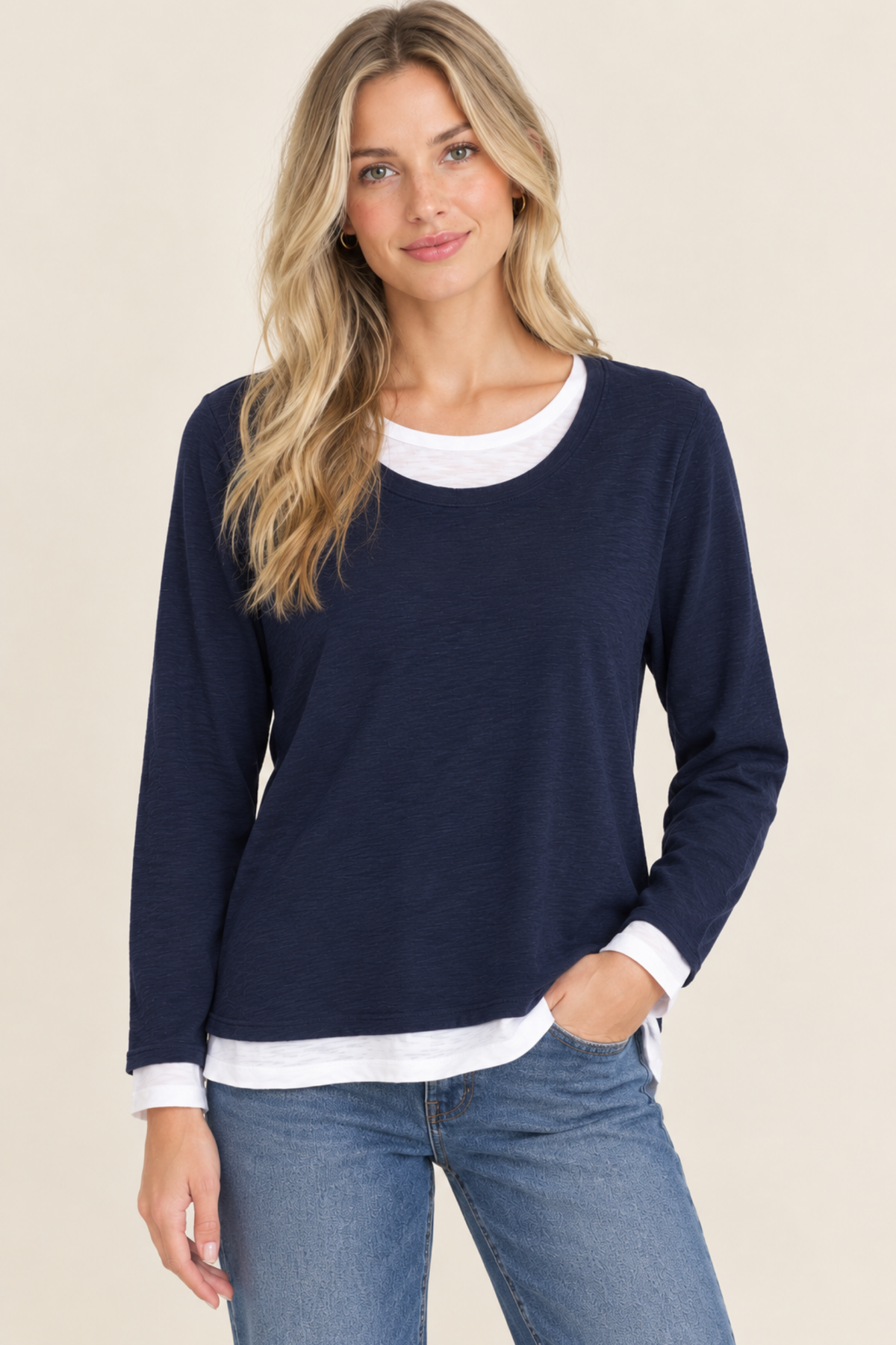 A woman modeling a navy blue long-sleeve crewneck sweater with a white layered crewneck underneath, paired with blue jeans against a solid beige background.