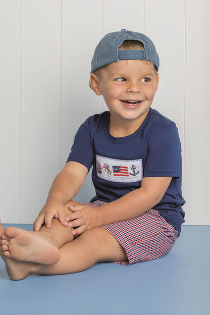 Child wearing a navy blue shirt with a graphic design and red plaid shorts, sitting on a blue surface against a white wall.