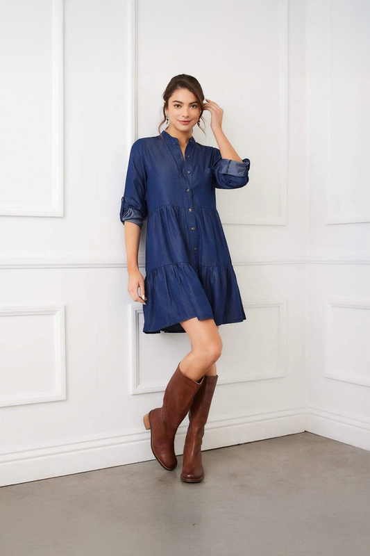 Woman wearing a blue dress and brown knee-high boots standing against a white paneled wall.