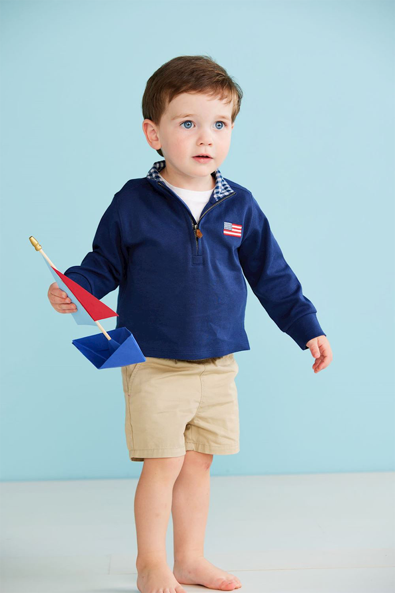 Child wearing a blue jacket with a flag patch, holding a toy boat against a light blue background