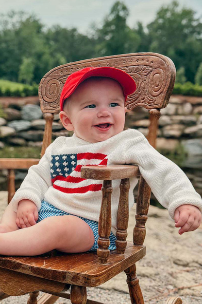 Baby sitting on a wooden chair wearing a red cap and a sweater with an American flag design.