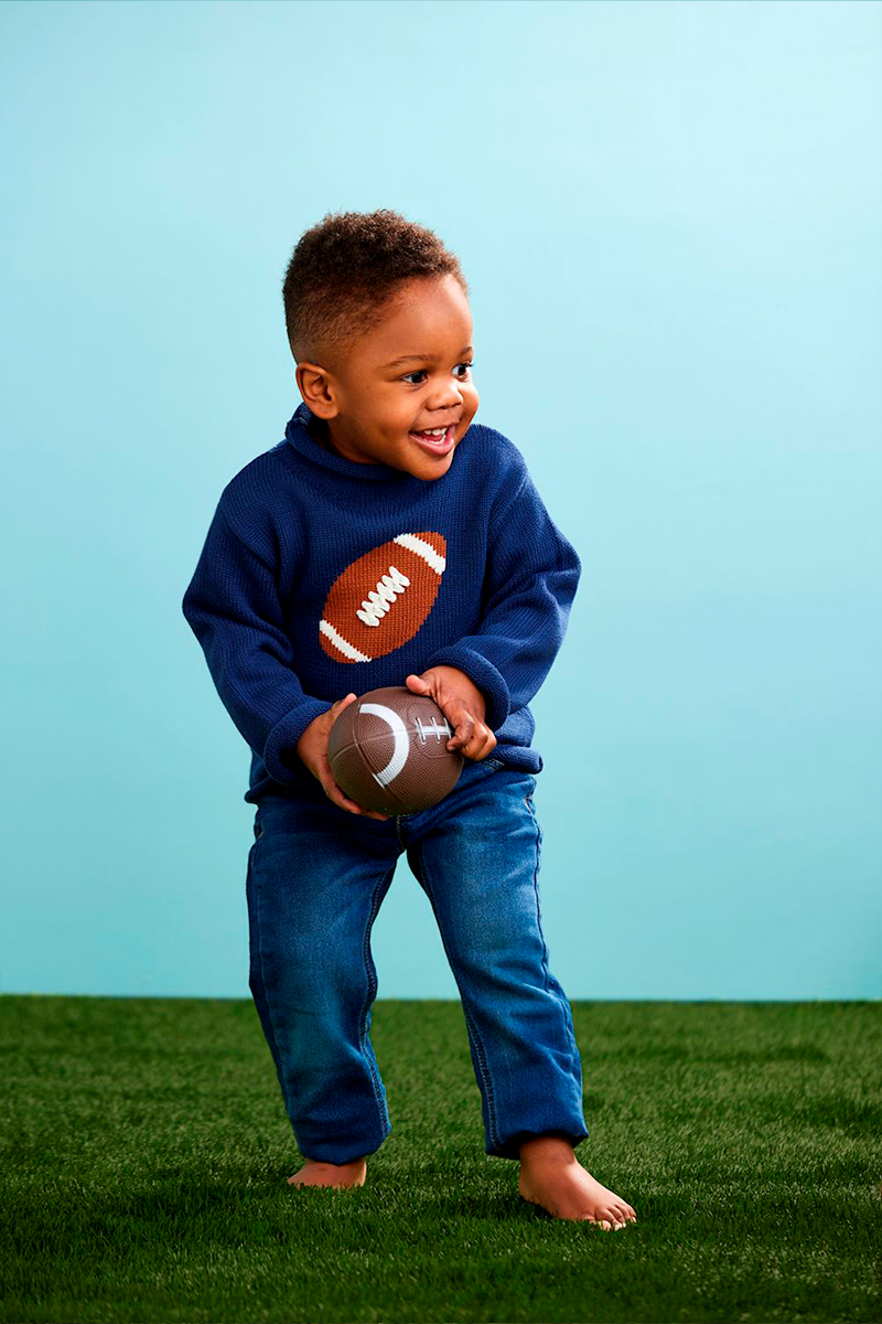 Child wearing a blue sweater with a football design, holding a football on a grassy field with a light blue background.