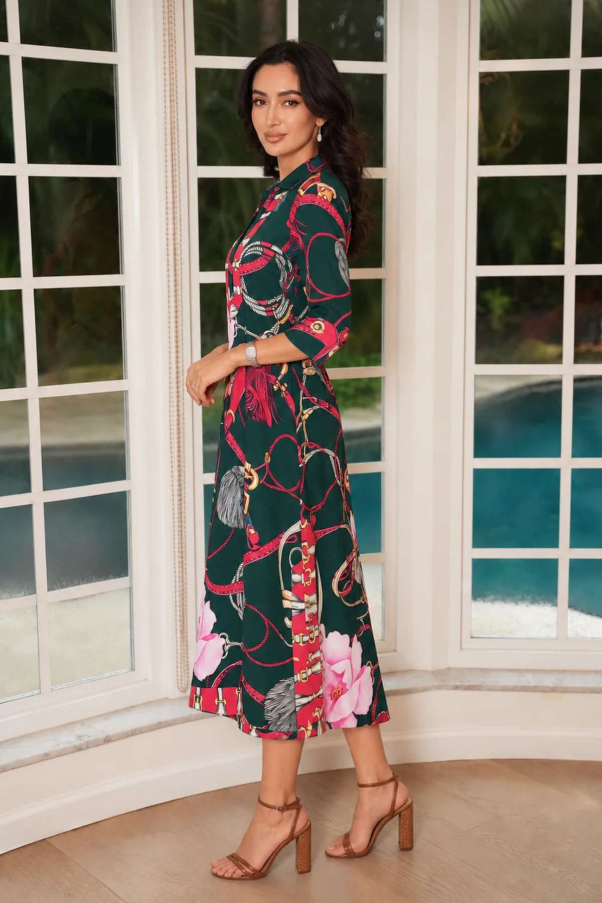 A woman modeling a dark green midi dress with a red, pink, and gold abstract floral print, standing in front of large white-framed windows with a pool visible outside.
