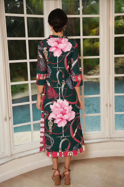 A woman viewed from behind wearing a dark green midi dress with a pink and red floral print, standing in front of a large white-framed window overlooking a pool.