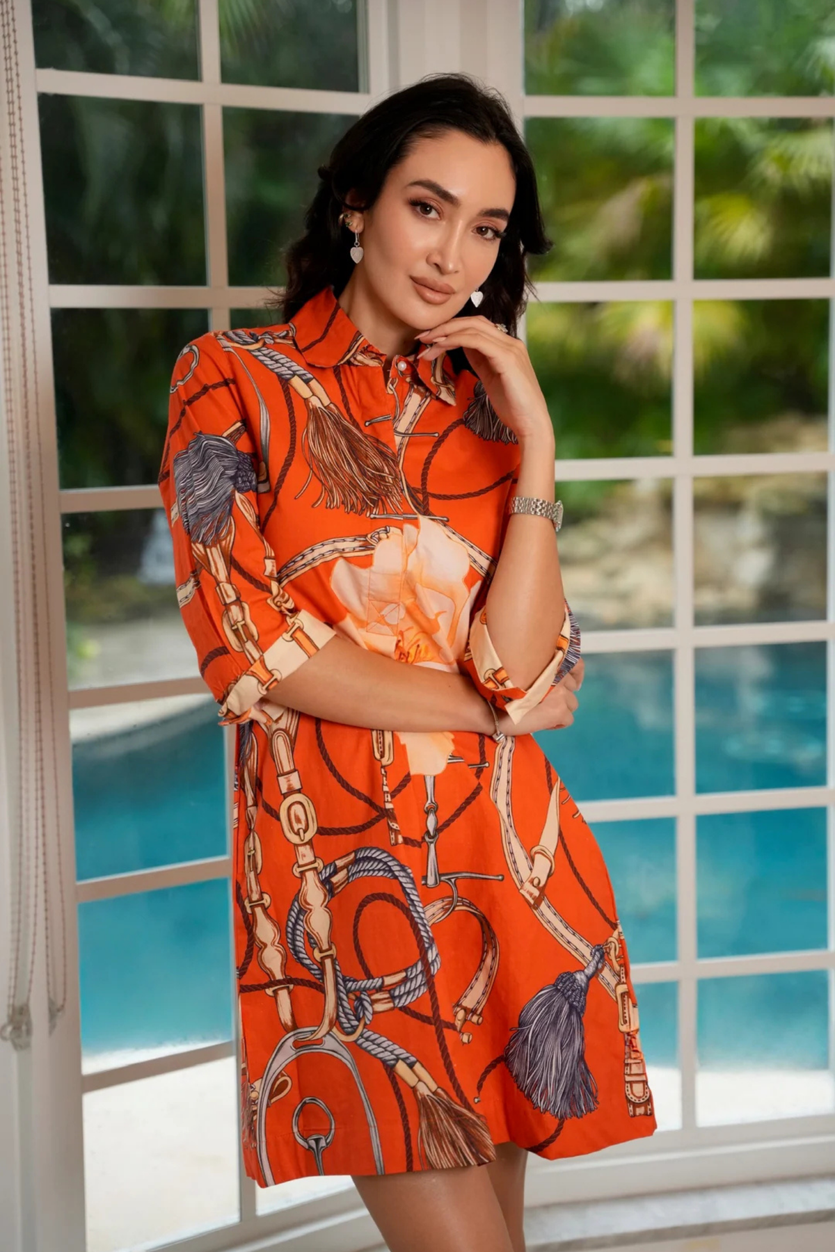 A woman models a short orange dress with a horse-bit and tassel print, standing in front of a large white-framed window overlooking a swimming pool.