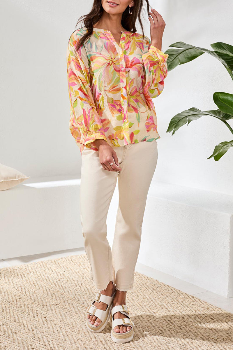 Woman wearing a colorful floral blouse and white pants in a bright room with a plant.