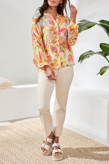 Woman wearing a colorful floral blouse and white pants in a bright room with a plant.