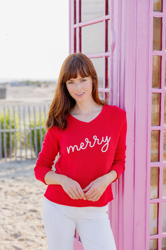 Woman wearing a red 'merry' sweater standing against a pink building on beach.