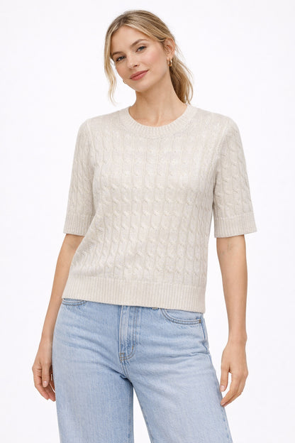 Woman wearing a cream metallic cable knit sweater and light blue jeans on a white background