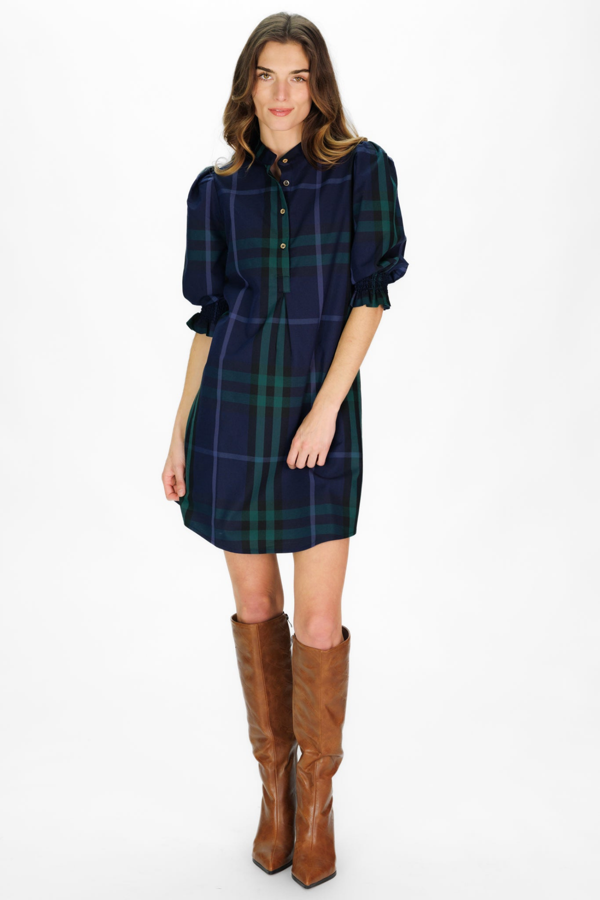 A woman modeling a navy blue tartan plaid shirt dress with green and red lines, featuring puff sleeves and a button-down front, paired with brown knee-high leather boots against a white studio background.