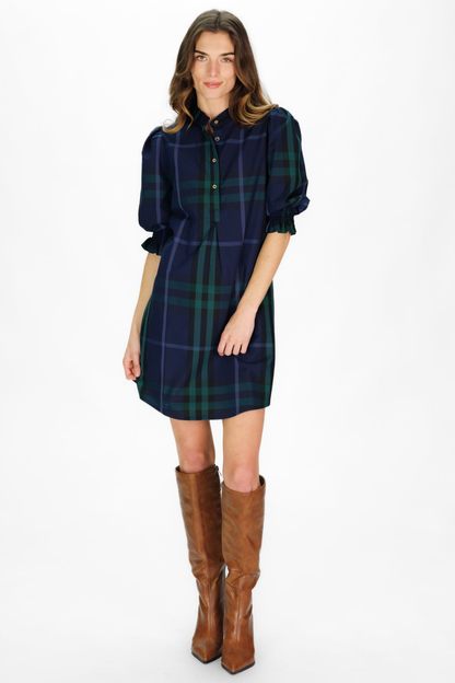 A woman modeling a navy blue tartan plaid shirt dress with green and red lines, featuring puff sleeves and a button-down front, paired with brown knee-high leather boots against a white studio background.