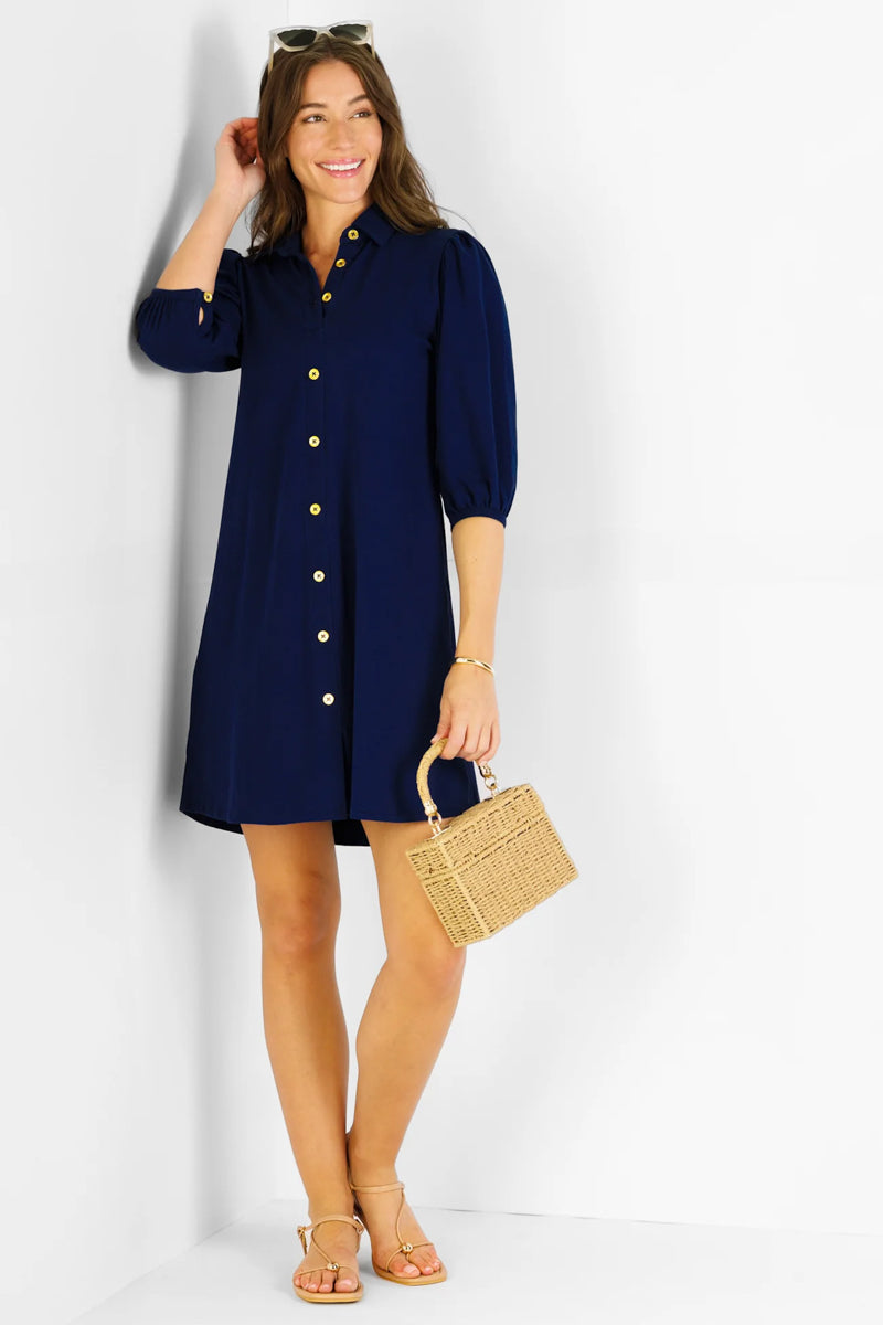 Woman wearing a navy blue dress with gold buttons, holding a straw bag on a white background