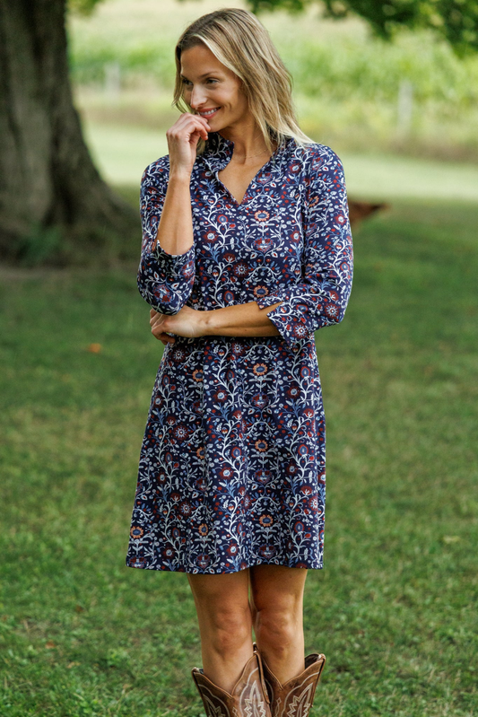 A woman wearing a navy blue floral print dress and brown cowboy boots, standing outdoors on a grassy lawn with a tree in the background.