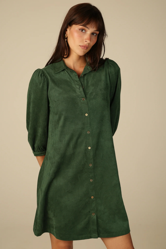 A woman wearing a dark green long-sleeve button-down shirt dress, standing against a plain beige studio background.