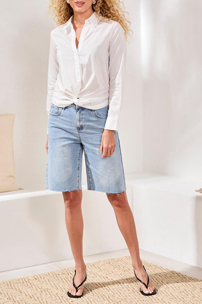 Woman wearing a white blouse and light blue denim shorts in a minimal indoor setting.