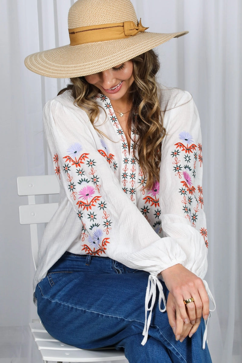 Woman wearing a white blouse with floral embroidery and a wide-brimmed hat, sitting on a white chair.