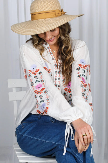Woman wearing a white blouse with floral embroidery and a wide-brimmed hat, sitting on a white chair.