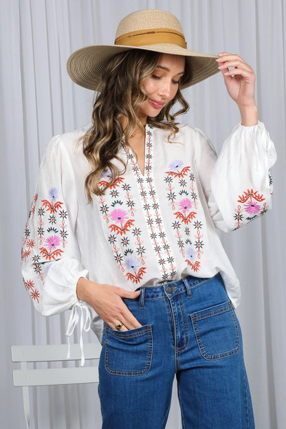 Woman wearing a white blouse with floral embroidery and blue jeans, holding a wide-brimmed hat.