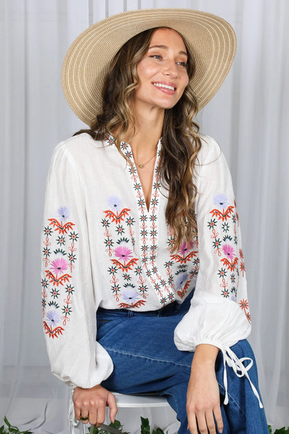 Woman wearing a white blouse with colorful embroidery and a wide-brimmed hat against a white curtain background.