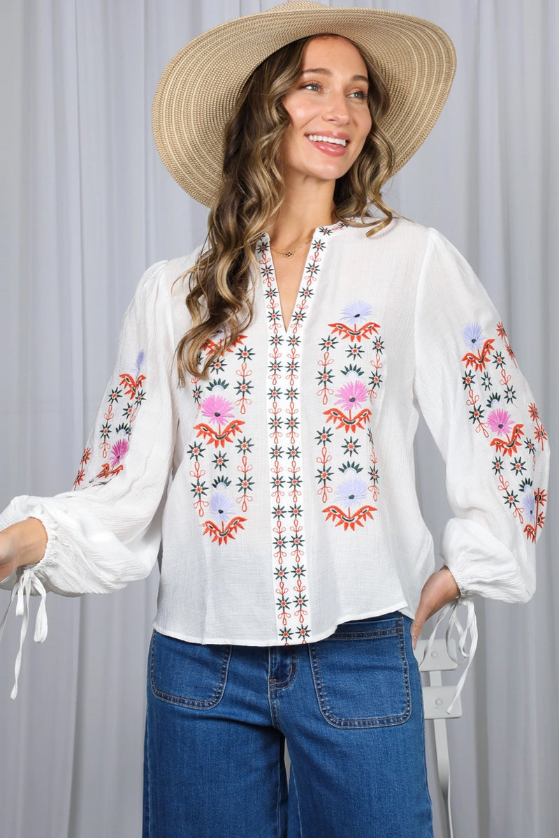 Woman wearing a white blouse with colorful embroidery and a wide-brimmed hat against a gray background
