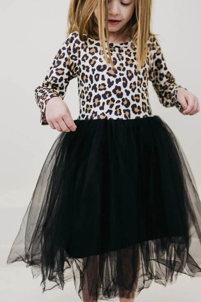 A young girl wearing a long-sleeve leopard print top and a black tulle tutu skirt, standing against a plain light background.
