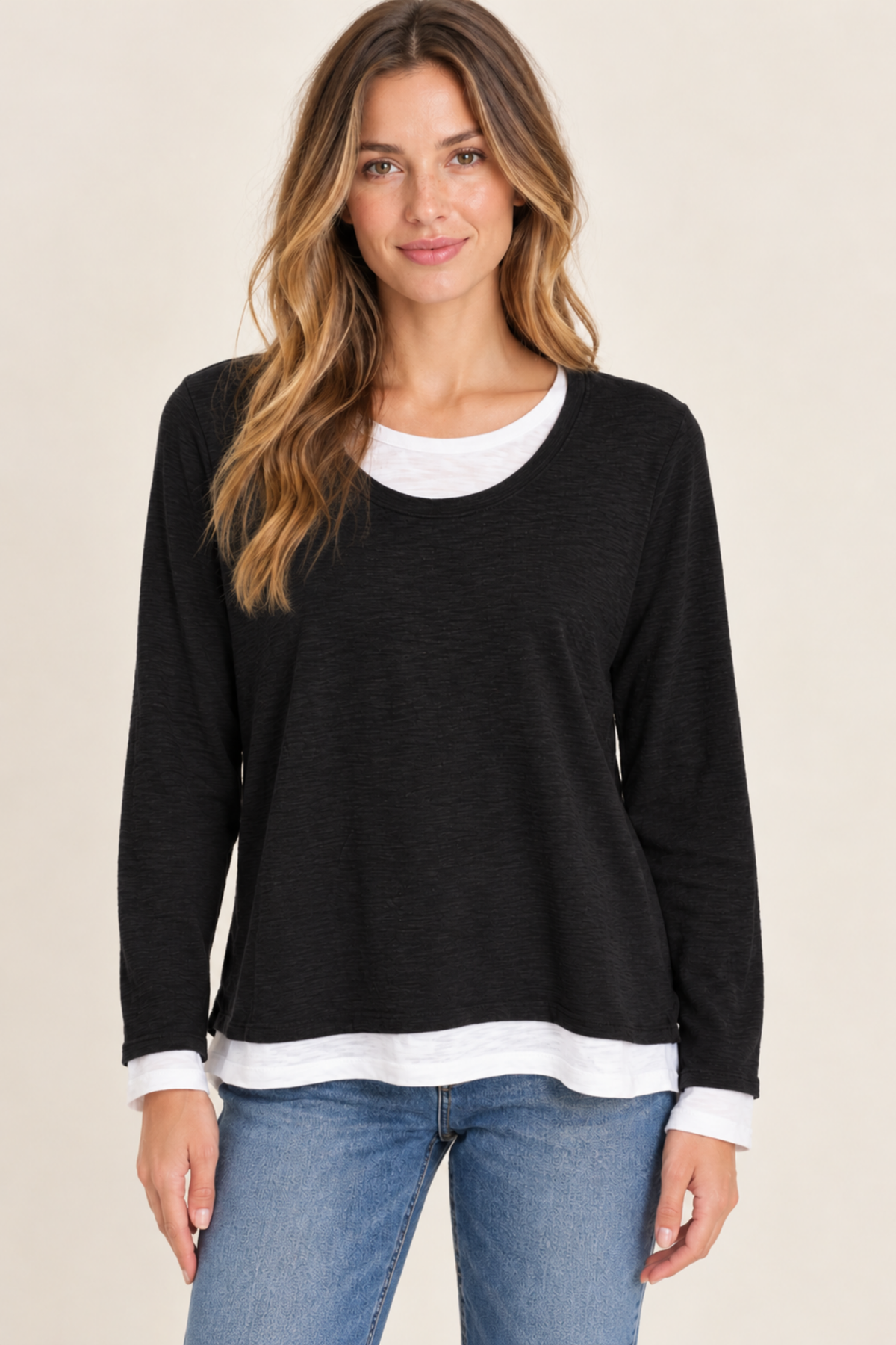 A woman wearing a black long-sleeve top with a white layered crewneck and hem detail, paired with blue jeans, against a plain beige background.